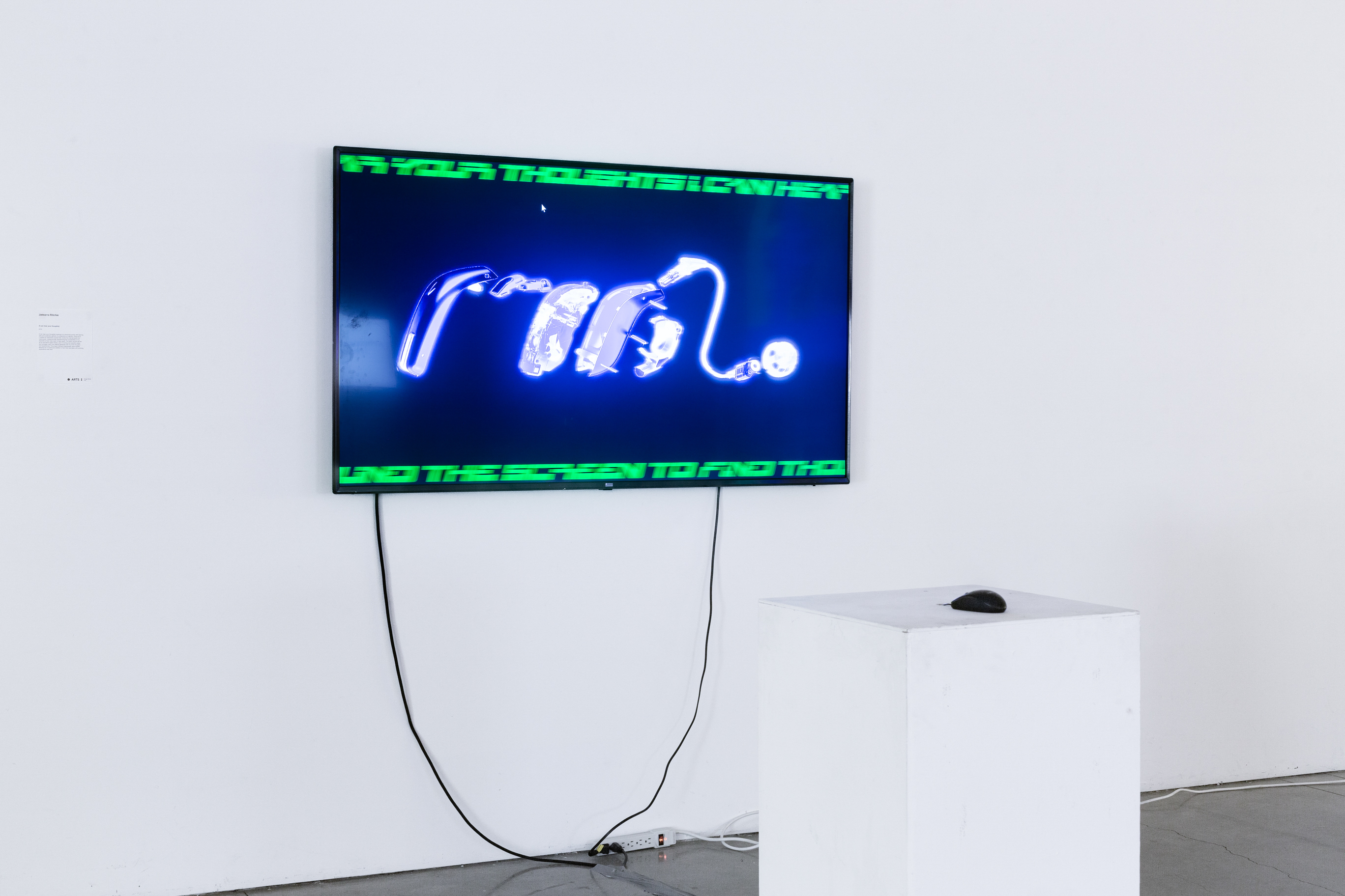 A TV mounted on a white wall, with visuals on it depicting hearing aids on a black background. In front of the TV is a podium with a computer mouse.